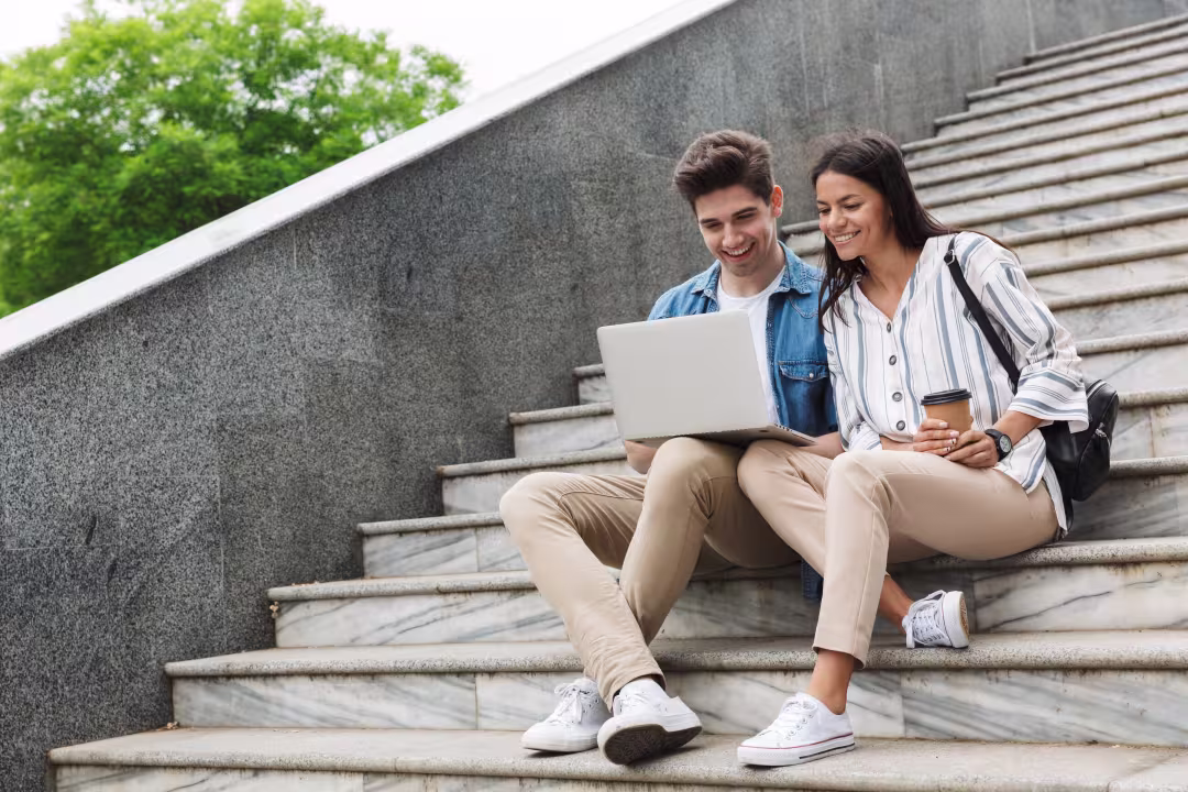 Zwei Personen sitzen gemeinsam auf einer Treppe und schauen auf den Bildschirm eines Laptops